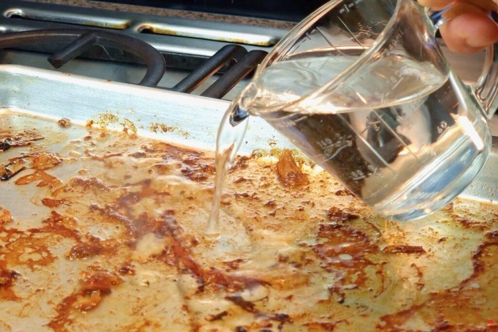 Water being poured from a jug into a roasting tin to loosen the browned bits