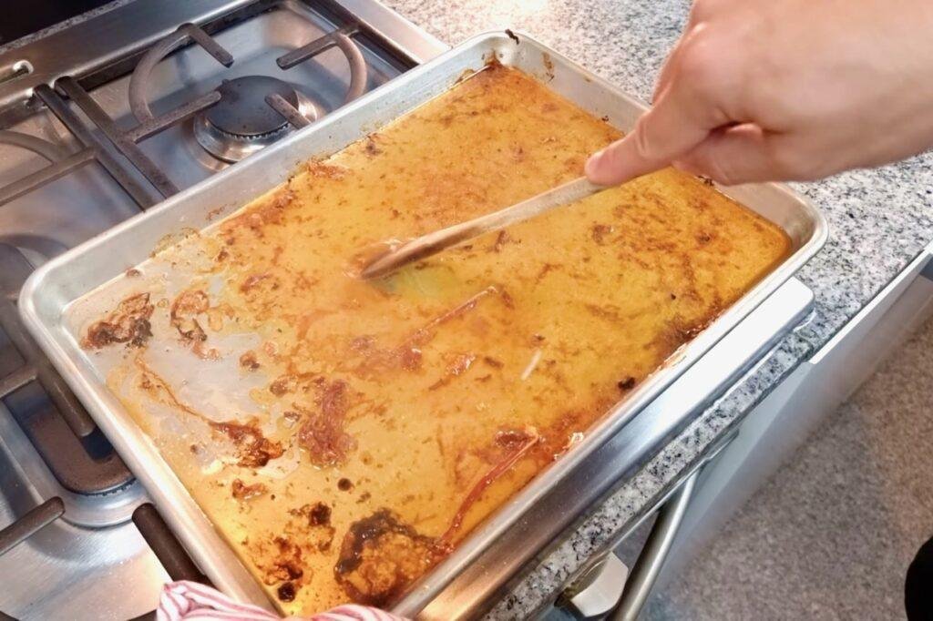 Hand scraping golden gravy in a metal tray with a wooden spoon