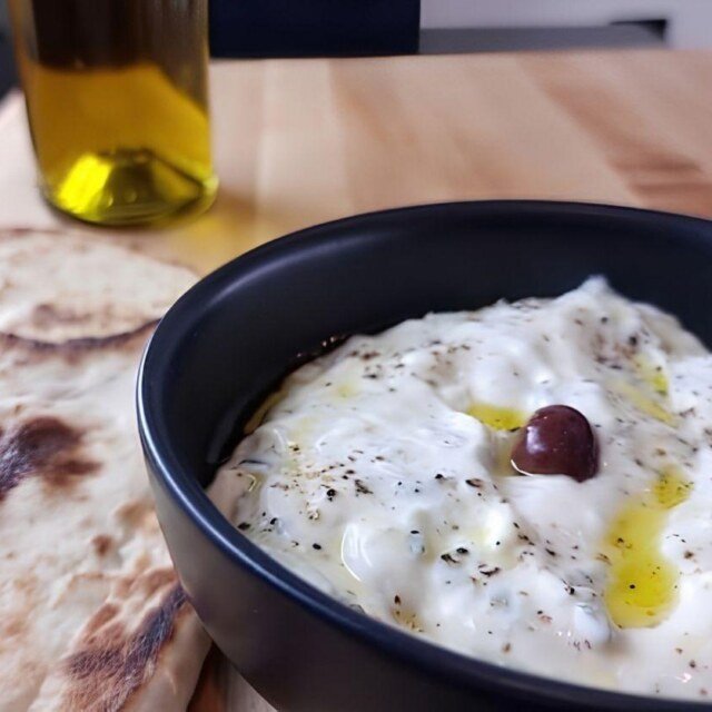 Creamy tzatziki in a bowl drizzled with olive oil, served with warm flatbreads and a bottle of olive oil in the background.