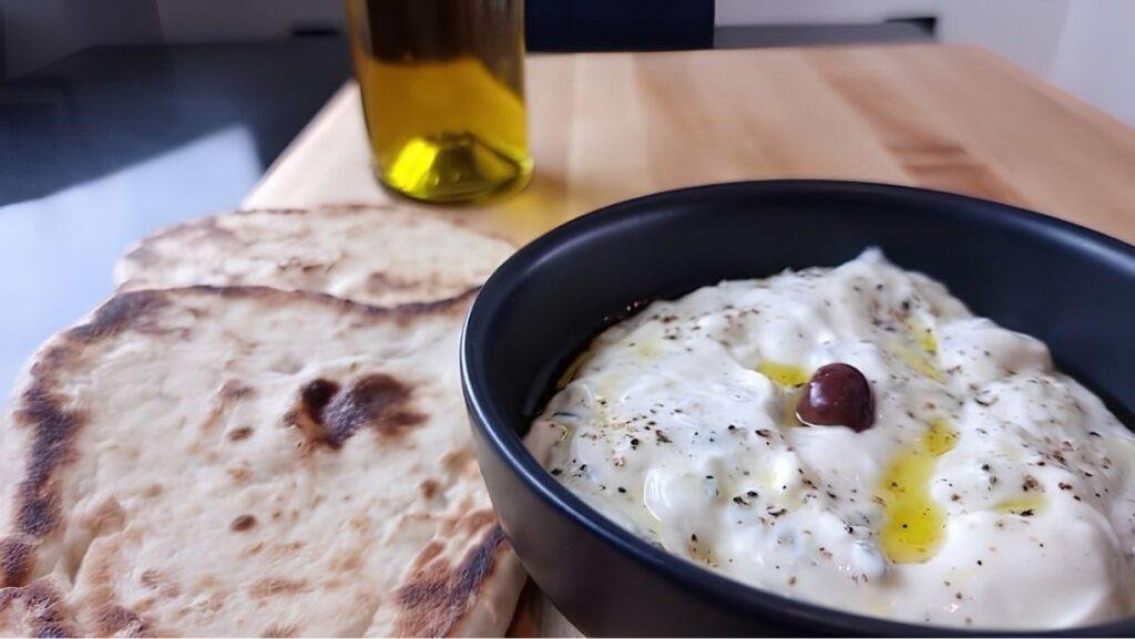 Creamy tzatziki in a bowl drizzled with olive oil, served with warm flatbreads and a bottle of olive oil in the background.