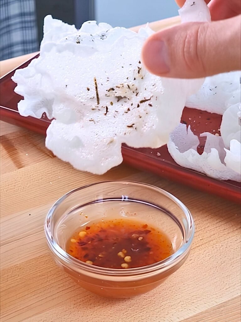 A hand holds a crispy vegan prawn cracker above a small bowl of dipping sauce, showcasing a tasty snack option.