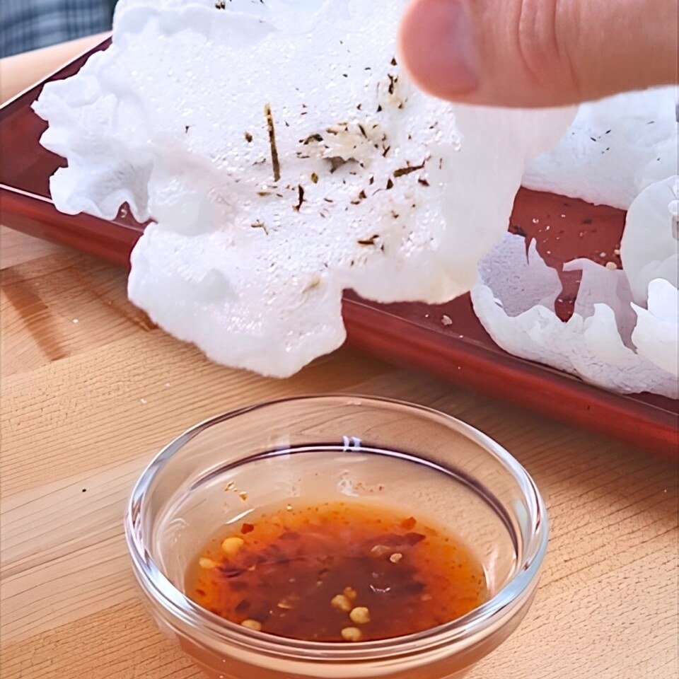 A hand holds a crispy vegan prawn cracker above a small bowl of dipping sauce, showcasing a tasty snack option.