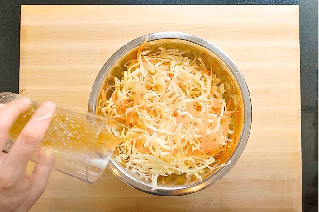 Hand pouring dressing over shredded cabbage and carrot in a steel bowl
