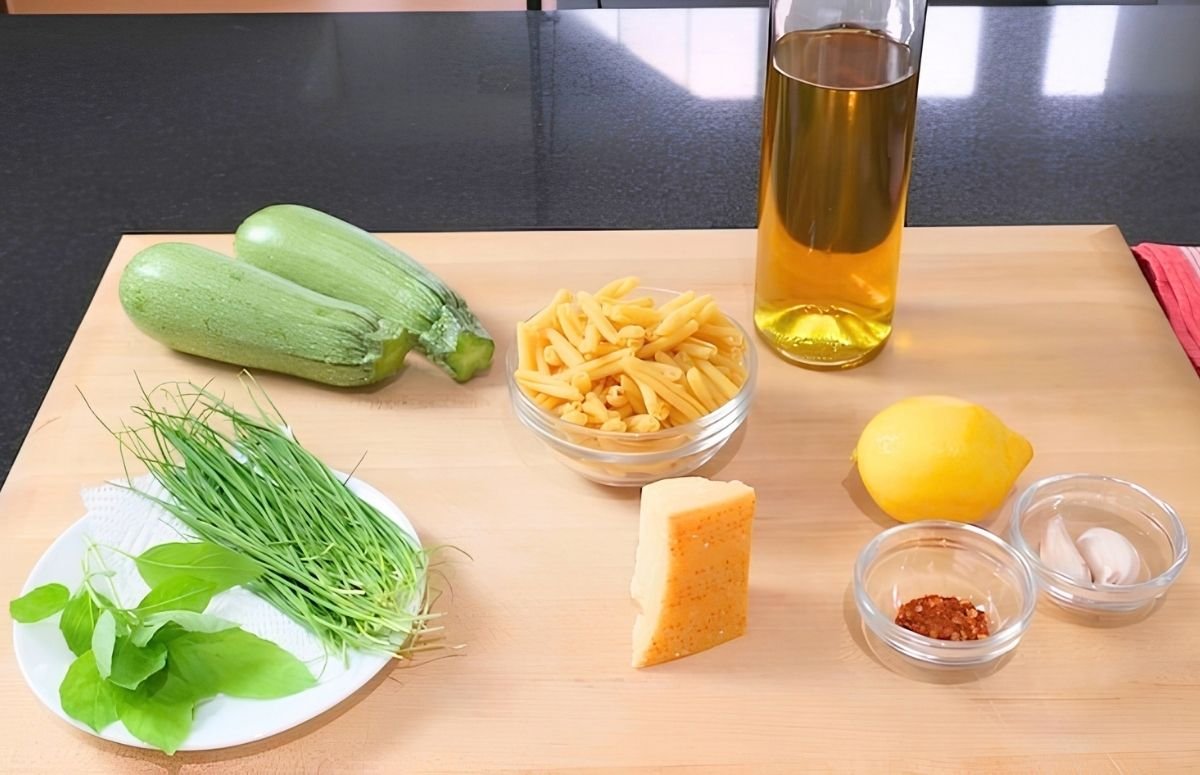 Ingredients laid out on a wooden board: courgettes, pasta, olive oil, lemon, parmesan, chives, basil, garlic and chilli flakes