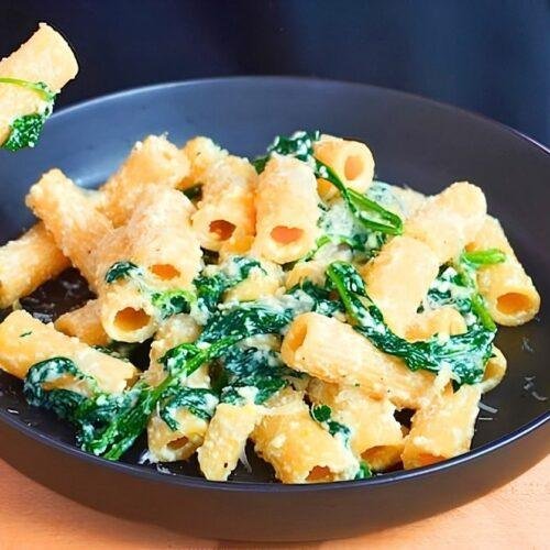 Fork lifting a piece of rigatoni from a dark bowl of spinach and ricotta pasta