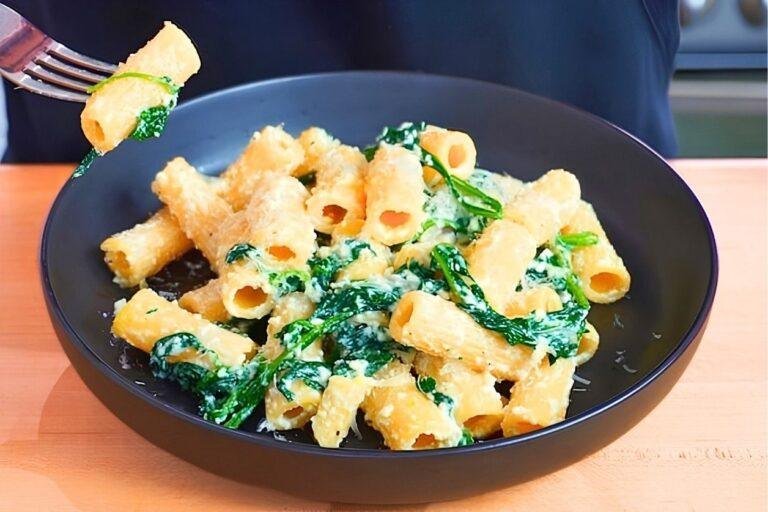 Fork lifting a piece of rigatoni from a dark bowl of spinach and ricotta pasta